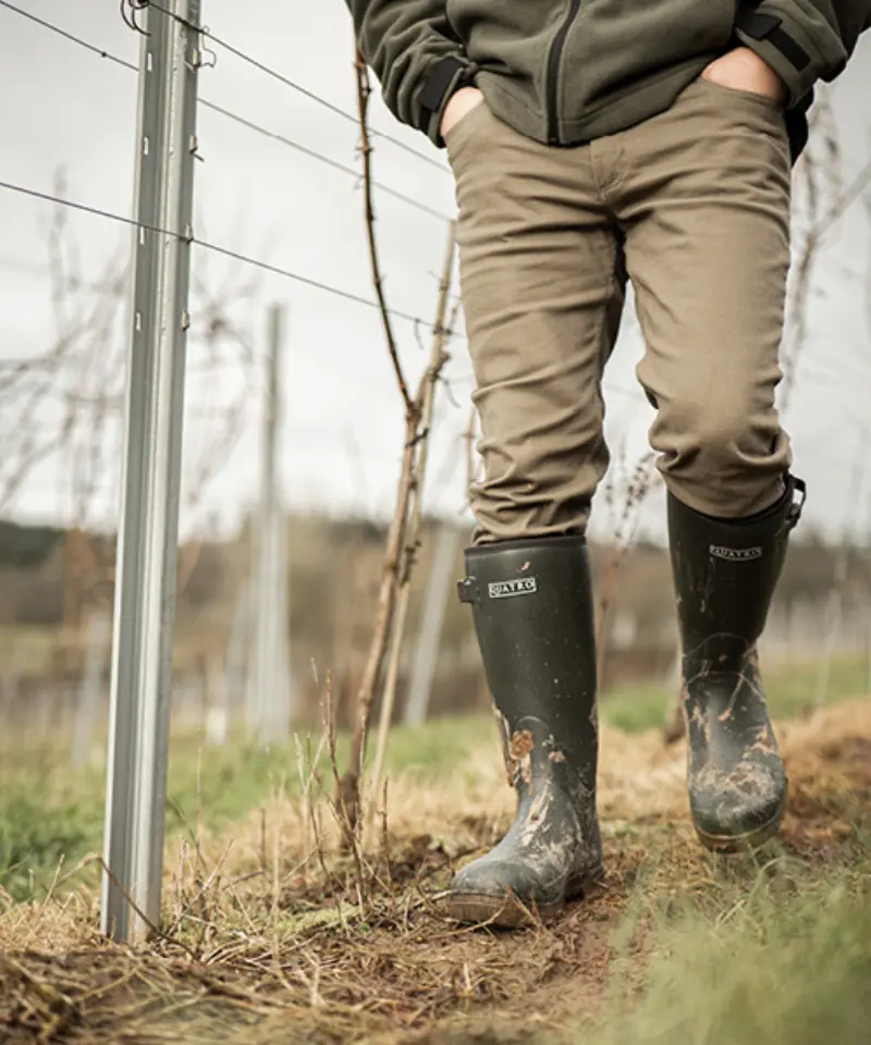 A person wearing muddy black rubber boots and khaki pants walks through a vineyard, with rows of bare grapevines and metal posts visible in the background.
